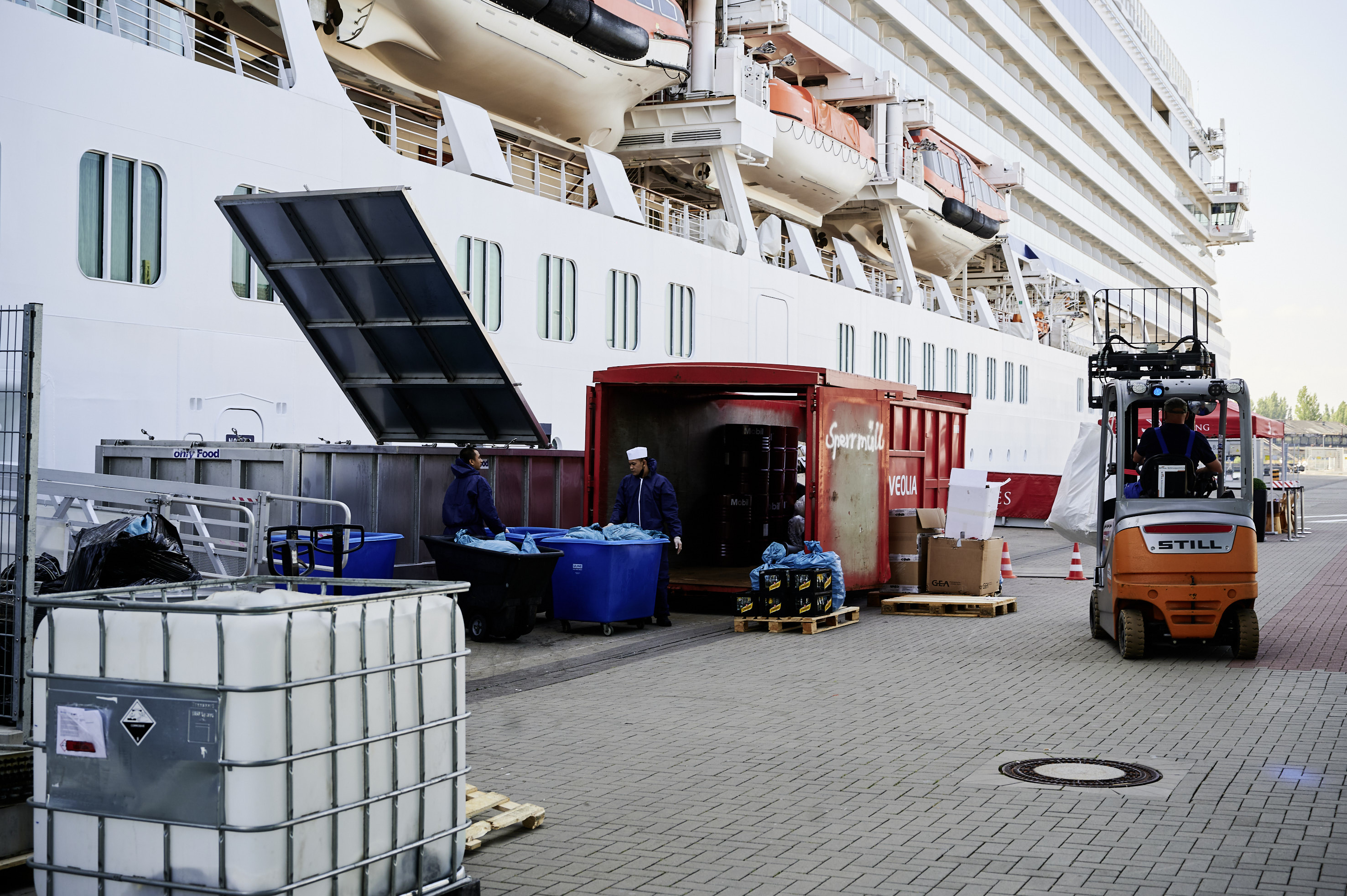 Kreuzfahrthafen Warnemünde: Zwei Personen schieben blaue, rollbare Boxen über die Pier, um Müll zu zwei Containern zu bringen. Ein Gabelstapler fährt vorbei, und im Hintergrund liegt ein großes Kreuzfahrtschiff.