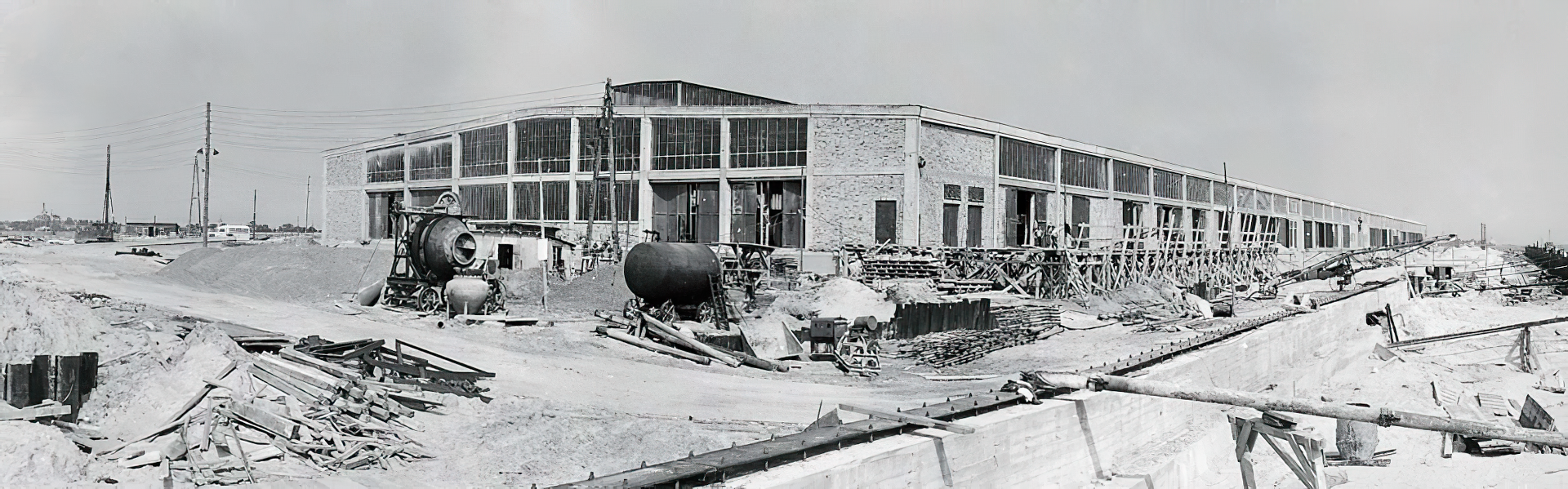 Historisches Schwarz-Weiß-Archivfoto: Baustelle einer großen Halle mit Betonmischern und Arbeitsgeräten im Vordergrund
