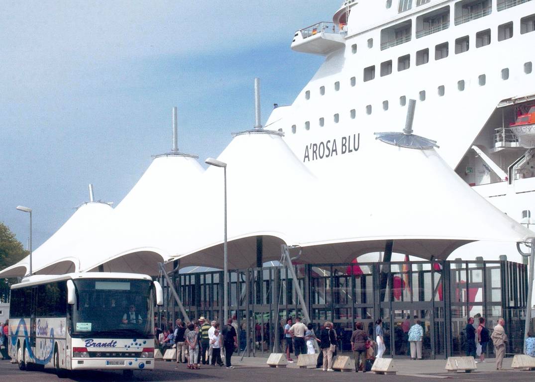 Archivbild des Kreuzfahrtterminals Warnemünde mit der gläsernen Abfertigungshalle auf Liegeplatz P7 und dem Kreuzfahrtschiff A'Rosa Blu im Hintergrund.