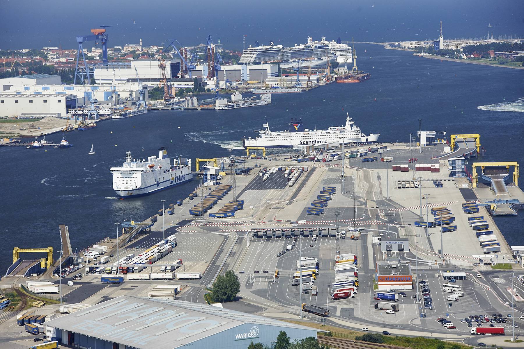 Luftbild des Fährterminals mit zwei Fähren im Hafen sowie dem Kreuzfahrtterminal mit zwei Kreuzfahrtschiffen im Hintergrund.