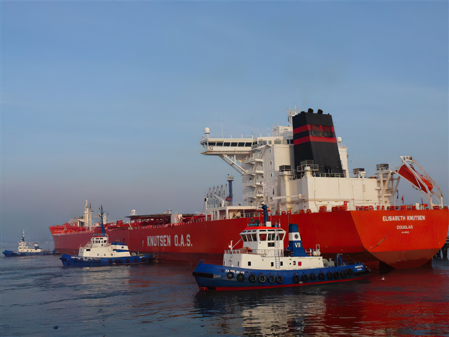 Ein großer roter Tanker im Ölhafen, der von drei Schleppern sicher an die Pier manövriert wird.