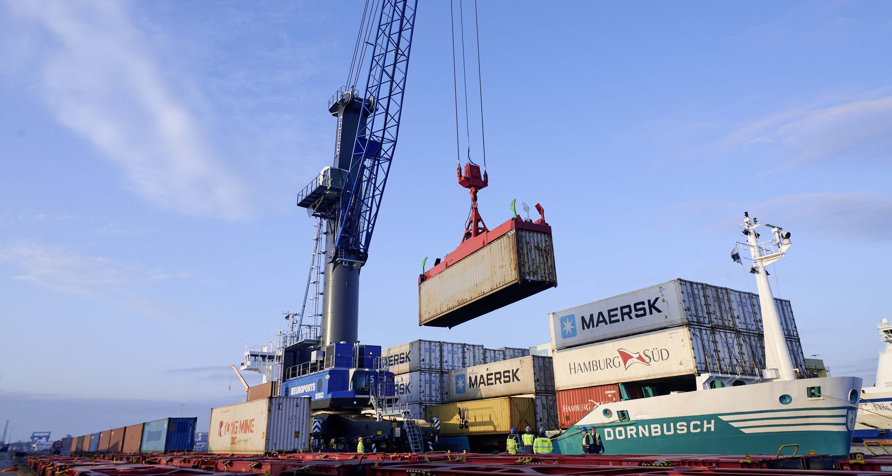 Ein Euroports-Hafenmobilkran beim Verladen eines Containers auf ein Schiff, fotografiert von der Landseite.