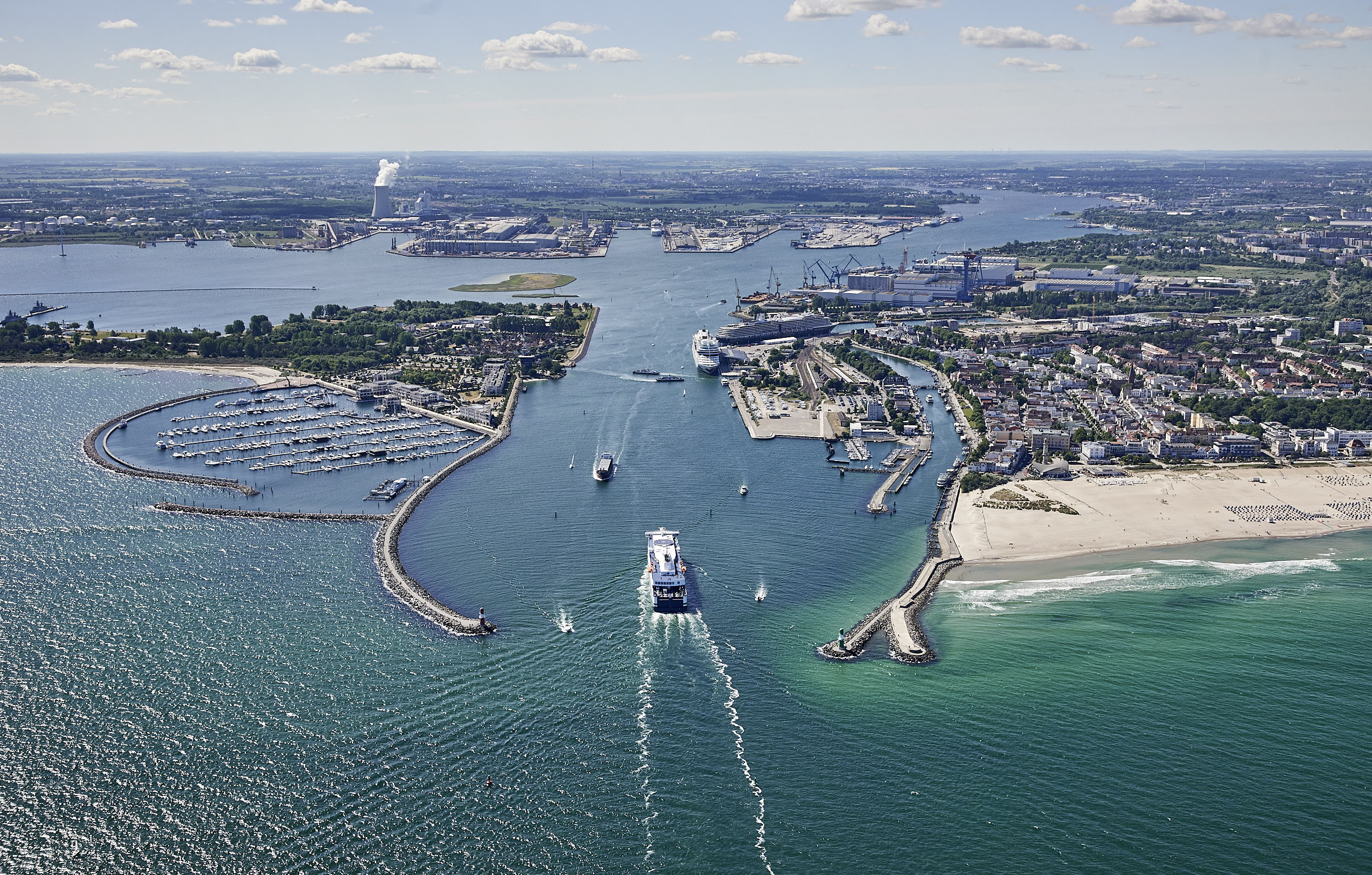 Luftaufnahme der Molenköpfe in Warnemünde. Eine Fähre fährt durch die Hafeneinfahrt. Zwei Kreuzfahrtschiffe liegen am Kreuzfahrtterminal in Warnemünde. Im Hintergrund ist der Rostocker Seehafen zu sehen.