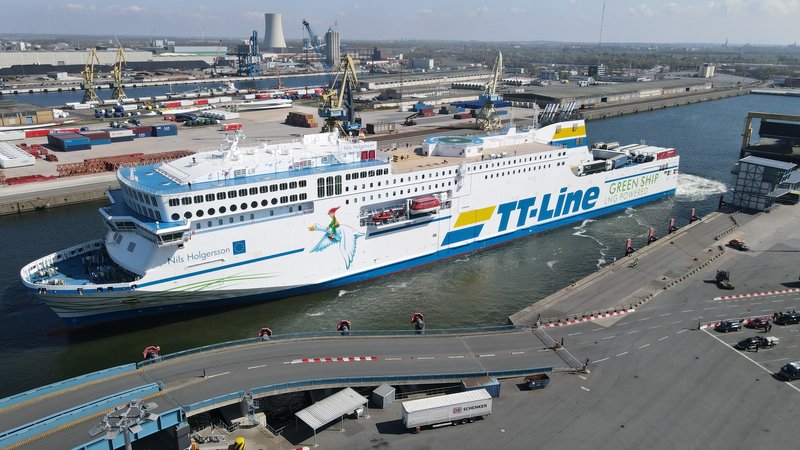 Luftaufnahme eines Fährschiffes im Hafenbecken. Im Hintergrund Industrie- und Logistikflächen vom Überseehafen Rostock. Vorne fahren Fahrzeuge auf ein weitere Autofähre.