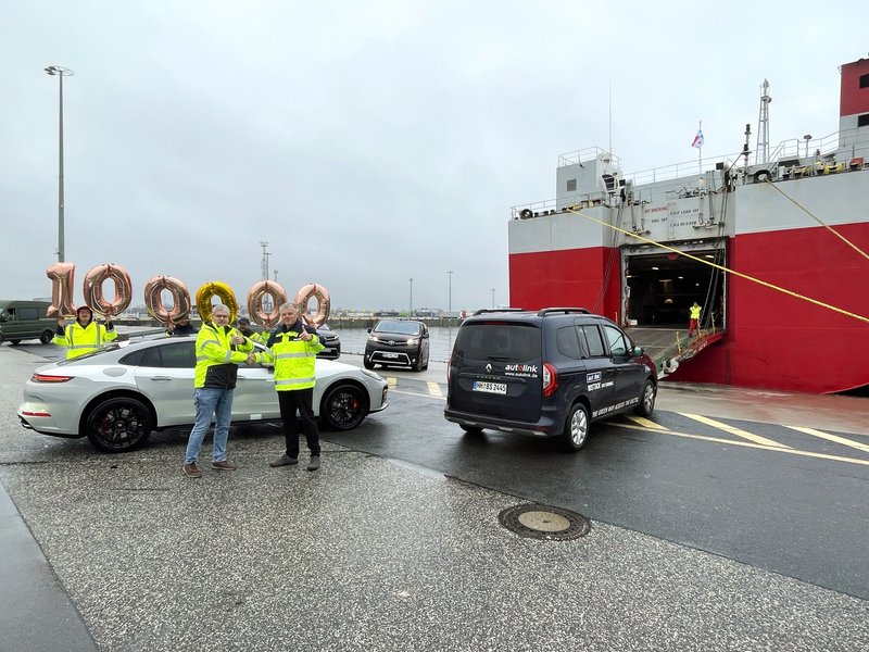 Jubiläum auf dem Autoterminal im Seehafen Rostock: Zwei Personen im Vordergrund schütteln sich lächelnd die Hände und zeigen den Daumen nach oben. Hinter ihnen bilden Luftballons die Zahl „100.000“, während im Hintergrund ein Autotransporter (Car Carrier) und drei verladebereite PKW zu sehen sind.