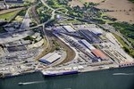 Luftaufnahme vom Terminal für kombinierten Ladungsverkehr im Überseehafen Rostock. Zwei Ganzzüge stehen auf den Gleisen unter den Hafenportalkränen, damit die Trailer verladen werden. Auf der Vorstellfläche stehen jede Menge Trailer. Eine RoRo-Fähre liegt am Liegeplatz. Auf der Warnow fährt ein Fahrgastschiff am Seehafen entlang.