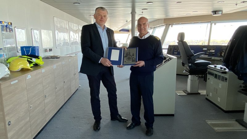 Zwei Männer auf der Kommandobrücke eines Schiffes überreichen sich eine Plakette.
