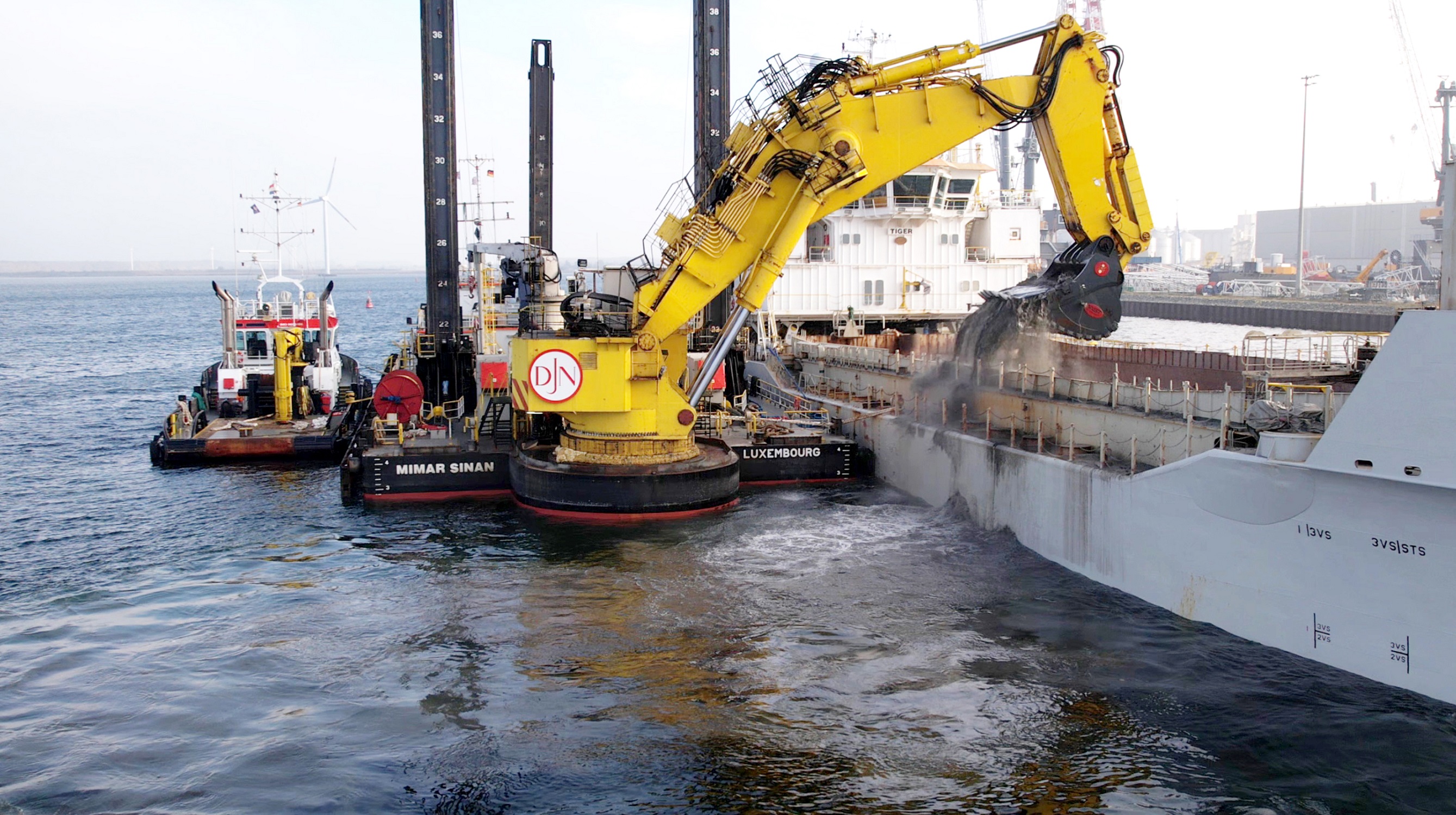 Seekanalvertiefung Rostock. Ein großer gelber Bagger der Firma Jan De Nul (DJN) auf einer schwimmenden Arbeitsplattform belädt ein Transportschiff auf dem Wasser mit Baggergut.