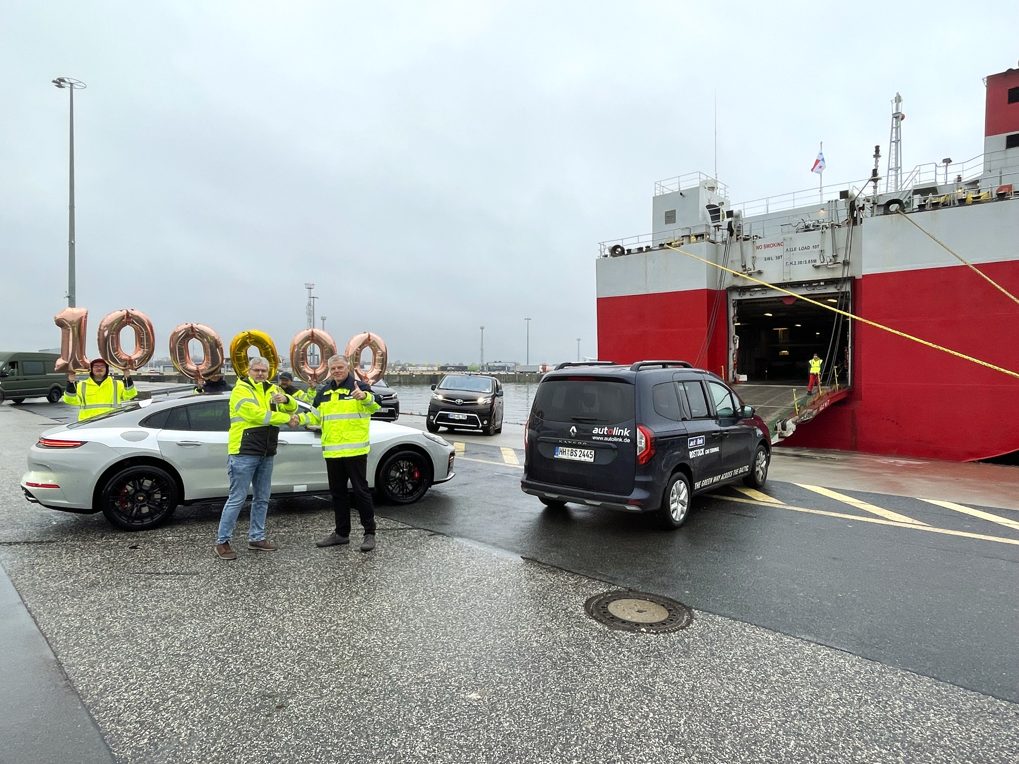 Jubiläum auf dem Autoterminal im Seehafen Rostock: Zwei Personen im Vordergrund schütteln sich lächelnd die Hände und zeigen den Daumen nach oben. Hinter ihnen bilden Luftballons die Zahl „100.000“, während im Hintergrund ein Autotransporter (Car Carrier) und drei verladebereite PKW zu sehen sind.
