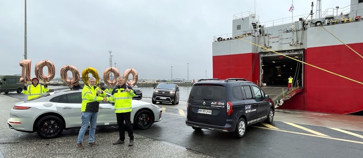 Jubiläum auf dem Autoterminal im Seehafen Rostock: Zwei Personen im Vordergrund schütteln sich lächelnd die Hände und zeigen den Daumen nach oben. Hinter ihnen bilden Luftballons die Zahl „100.000“, während im Hintergrund ein Autotransporter (Car Carrier) und drei verladebereite PKW zu sehen sind.