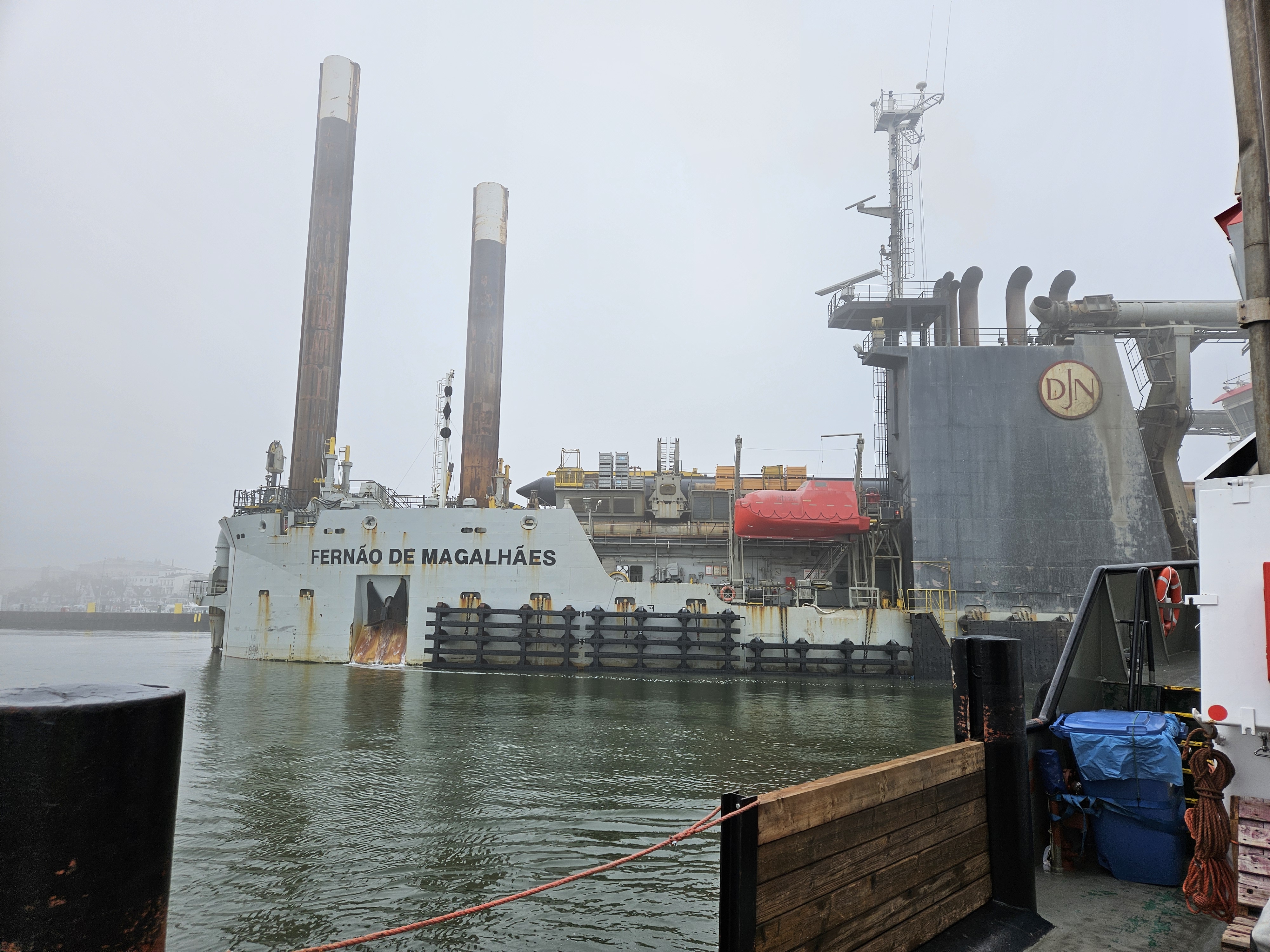 Ein großes, spezialisiertes Baggerschiff (Schneidkopfmutterbagger) namens „Fernão de Magalhães“ der Firma Jan De Nul (DJN) liegt bei nebligem Wetter im Seekanal Rostock. Markant sichtbar sind die beiden hoch aufragenden Verankerungspfähle (Spuds) und der Schiffsaufbau mit dem DJN-Logo.