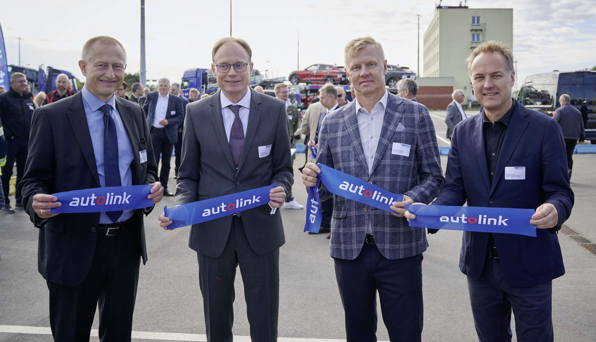 Gruppenaufnahme von vier Männern in Business-Kleidung, die auf dem Autoterminal im Seehafen Rostock stehen und lächelnd in die Kamera blicken. Sie halten jeweils ein Band mit der Aufschrift „autolink“ präsentierend nach vorne.