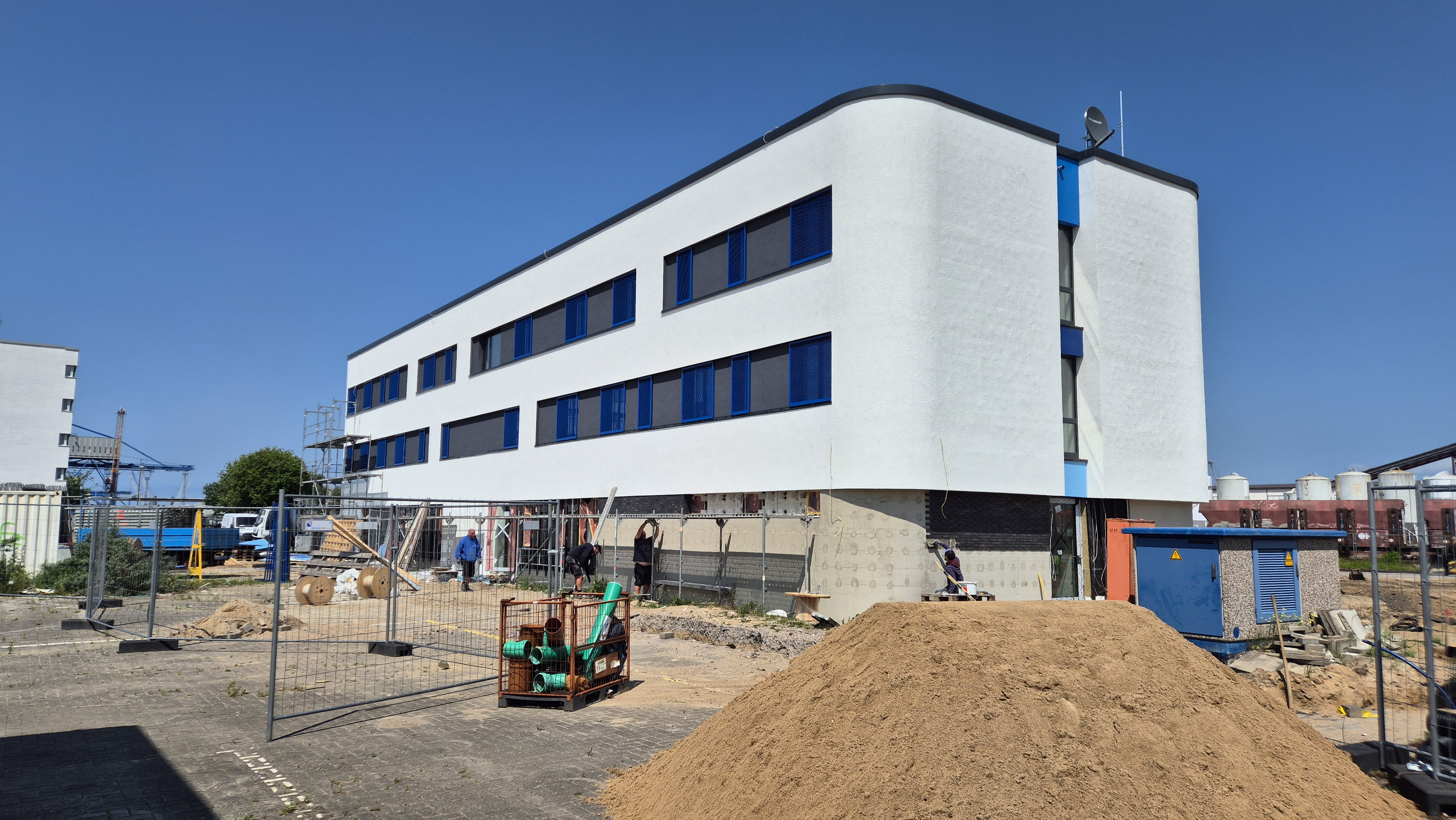 Außenaufnahme des neuen Bürogebäudes der Bundespolizei im Seehafen Rostock bei sonnigem Wetter. Die moderne Fassade ist nach dem weitgehenden Gerüstabbau bereits gut sichtbar; lediglich an der kurzen Stirnseite des Hauses steht noch ein verbleibendes Baugerüst.