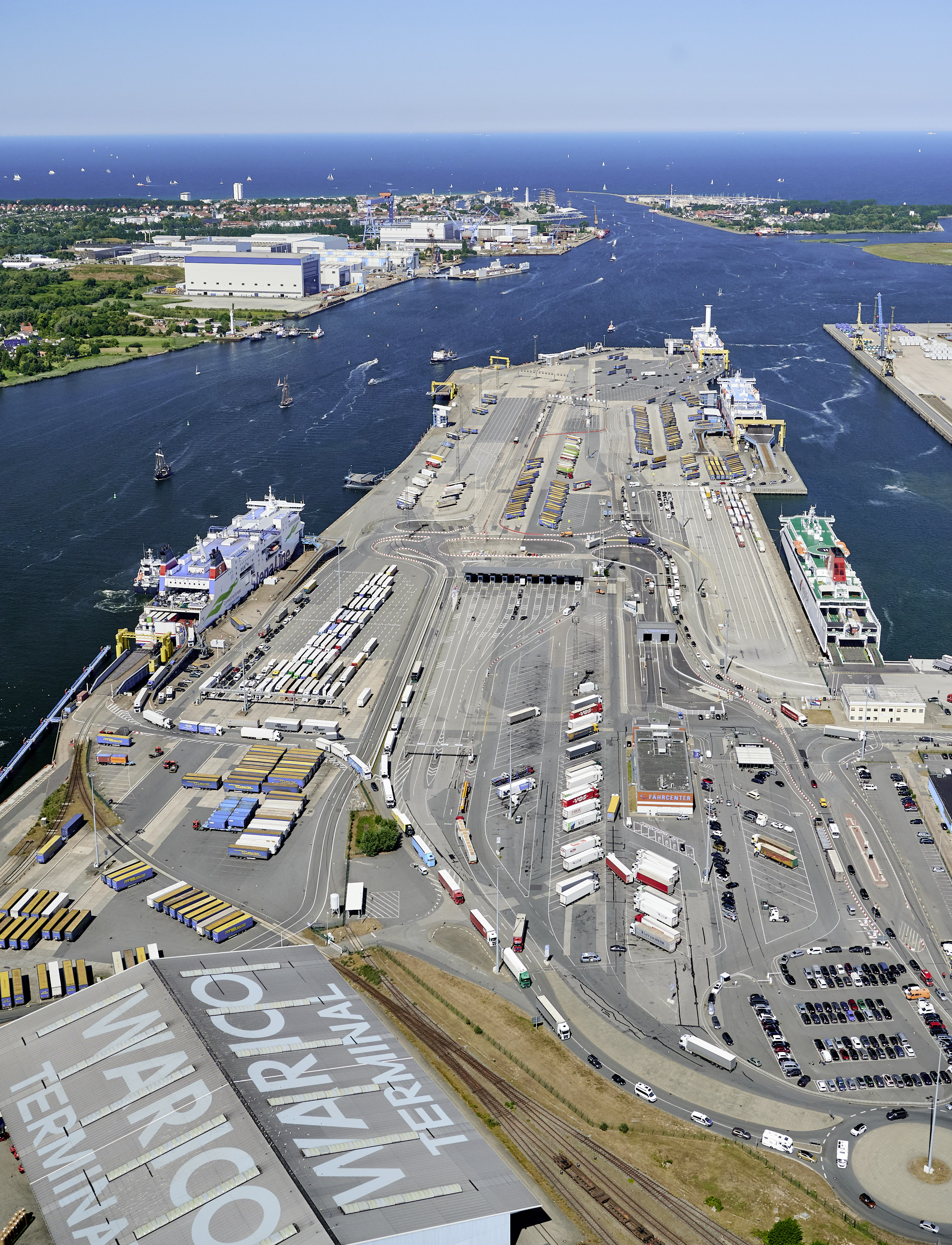 Aerial view of the ferry terminal at the Port of Rostock from the south. Trailers, rail wagons, and passenger cars are waiting in the staging areas to board the four ferries already moored alongside the quay.