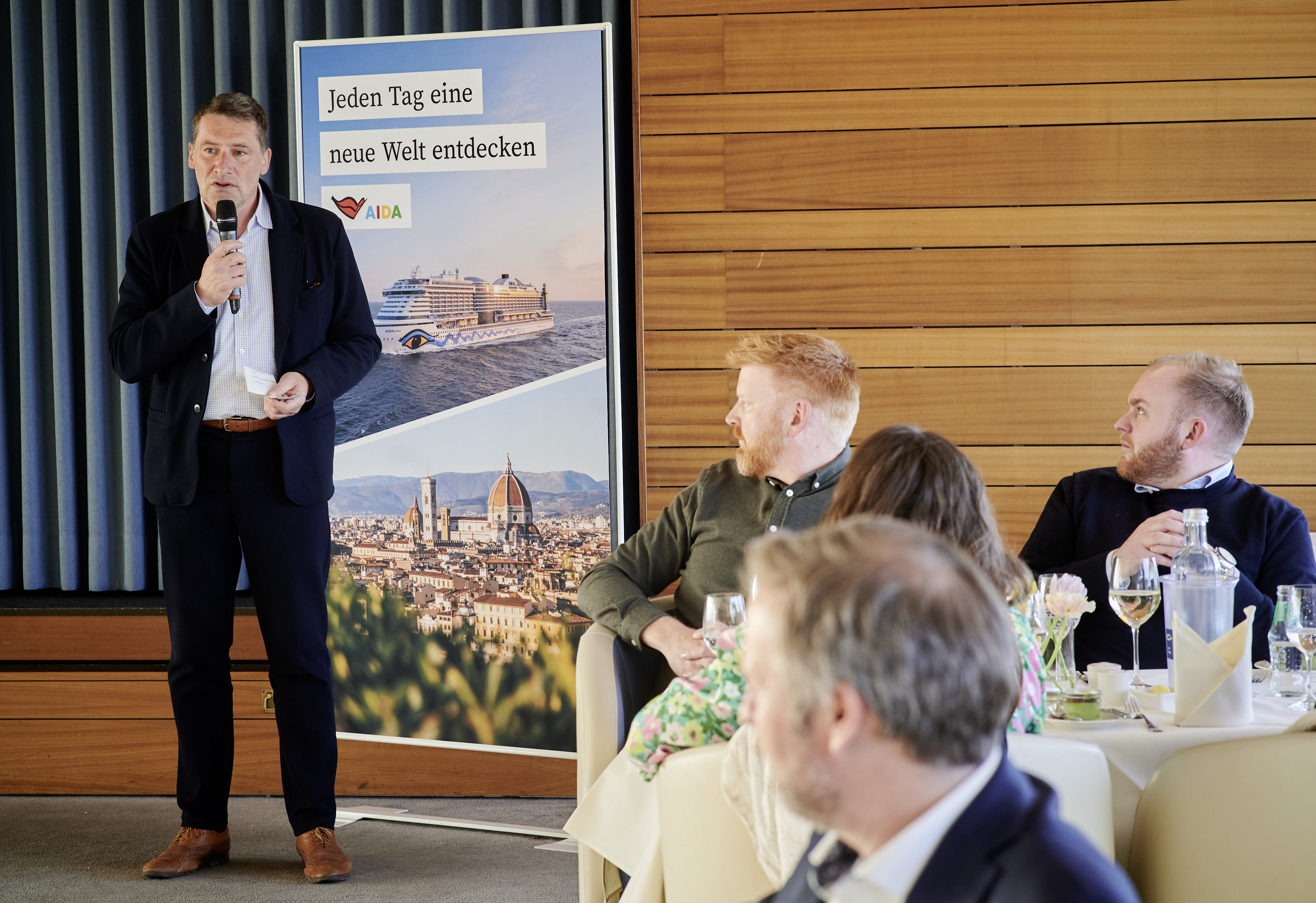 ROSTOCK PORT-Geschäftsführer Jens Scharner spricht in ein Mikrofon. Hinter ihm ein Roll-Up von AIDA.