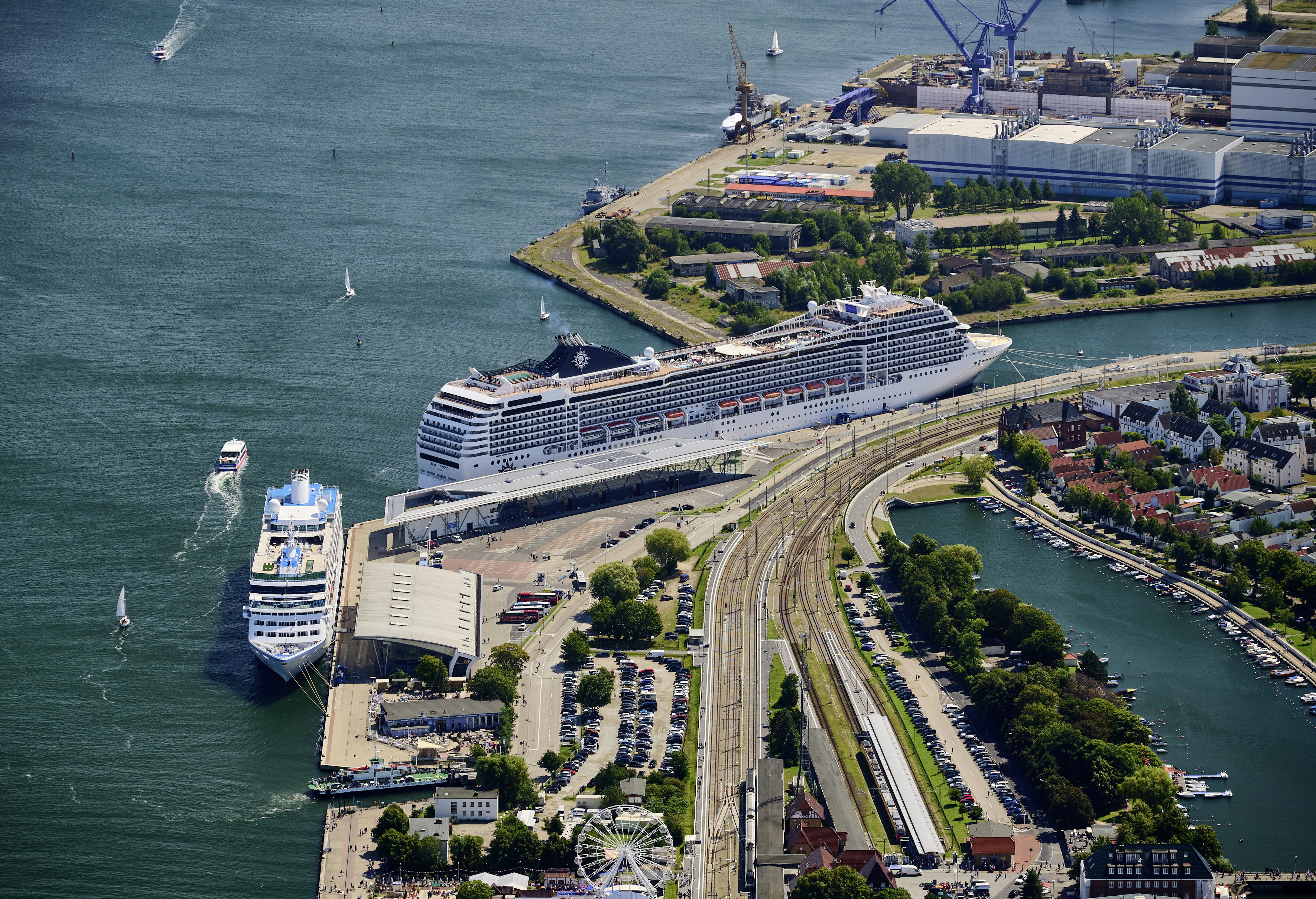 Luftaufnahme des Rostocker Überseehafens mit Blick über mehrere Hafenbecken bis zur Ostsee. Im Vordergrund verlaufen Straßen, Kreisverkehre und ausgedehnte Gleisanlagen, die den Hafen an das Verkehrsnetz anbinden. Dahinter erstrecken sich Kaianlagen, Lagerflächen, Kräne und Terminalgebäude. Mehrere Schiffe liegen an den Piers oder befinden sich auf der Wasserstraße. Im Hintergrund sind die Mündung zur Ostsee, Warnemünde sowie die Küstenlinie zu erkennen