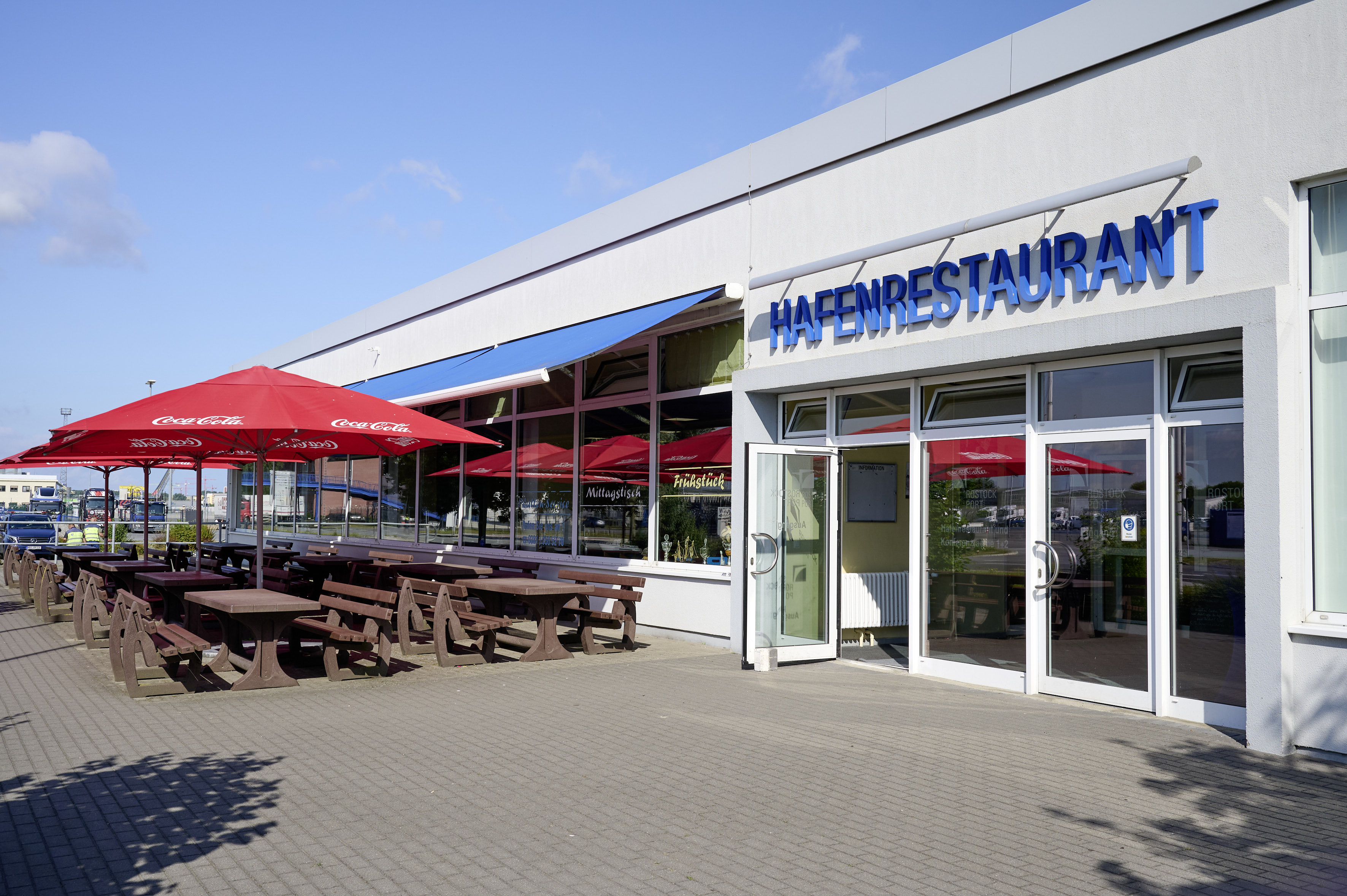 Eingang zum Hafenrestaurant im Überseehafen Rostock. Die Tür ist geöffnet. Über den Sitzplätzen im Außenbereich sind Sonnenschirme aufgespannt.